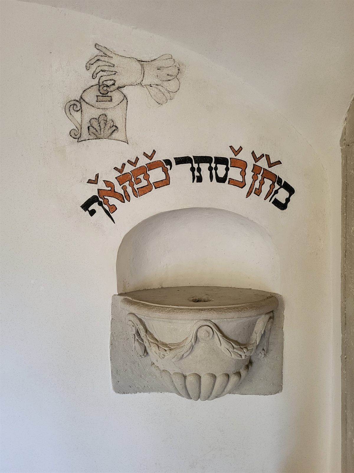 A carved stone charity box set into a whitewashed wall inside the Mád synagogue, with a painted hand dropping a coin into a box and a Hebrew inscription above it