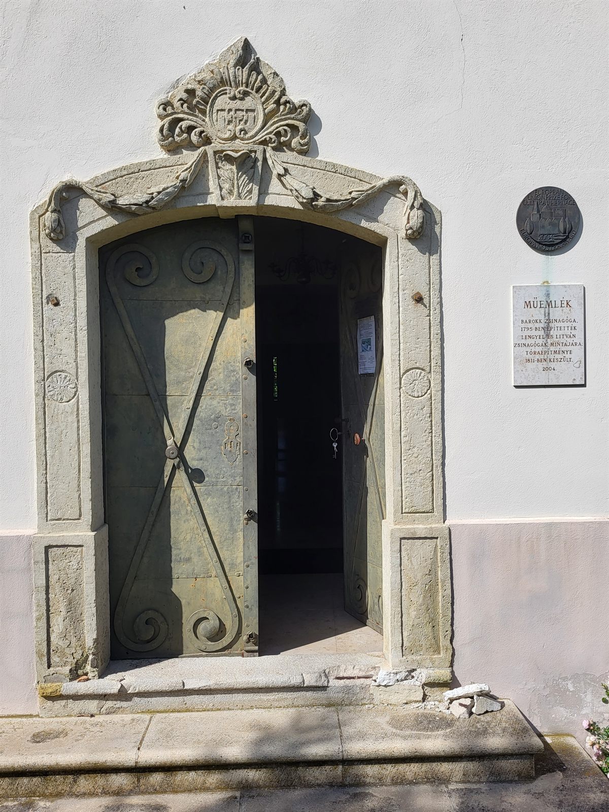 The carved stone entrance portal of the Mád synagogue, with a crowned cartouche above the doorway and the iron-bound wooden doors standing open