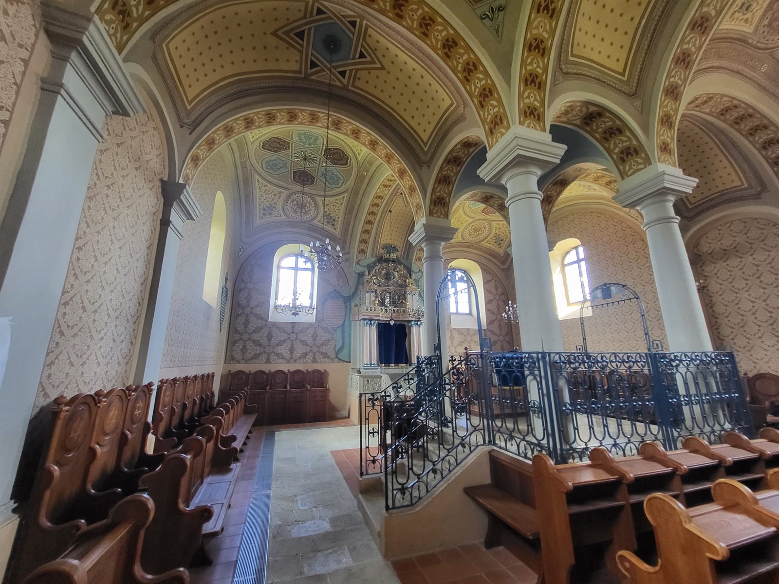 The vaulted prayer hall of the Mád synagogue seen from the side, with painted ceiling vaults, tall arched windows, wrought-iron railings around the bimah and rows of wooden benches