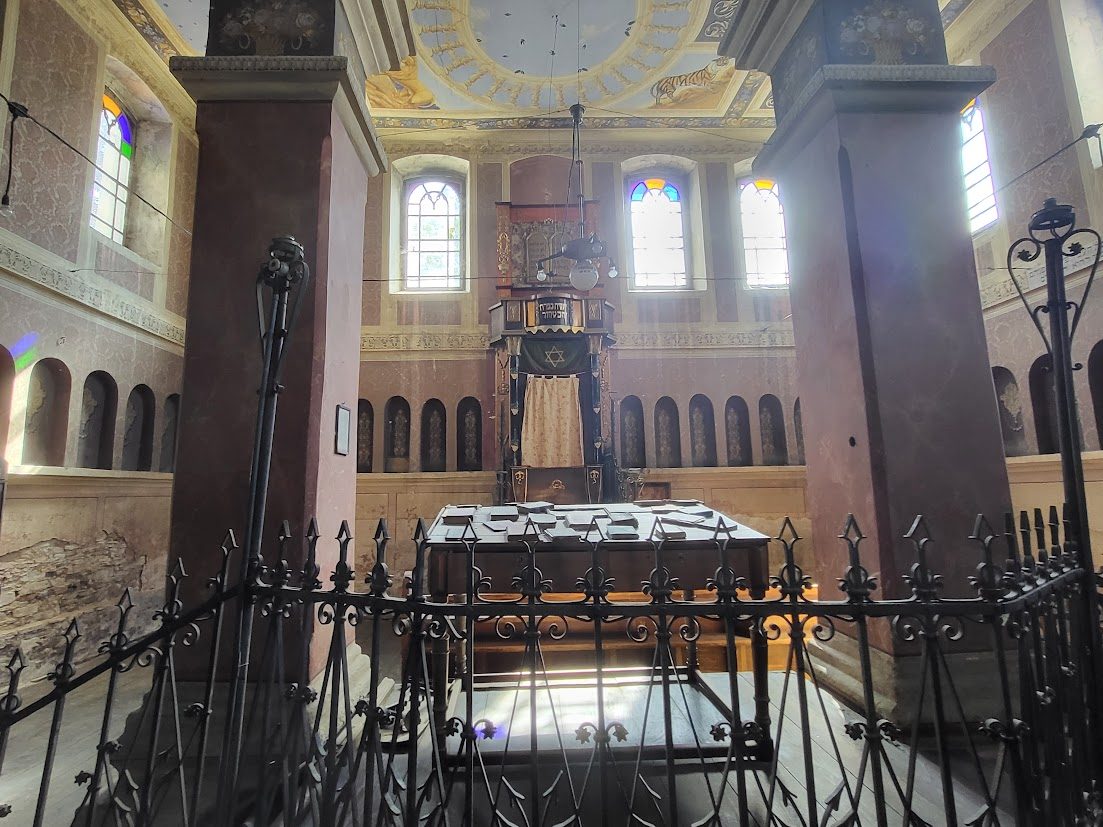 Interior of the Siret synagogue: the Torah ark between tall arched windows with stained glass, painted vaulted ceiling, and the bimah enclosed by a wrought-iron railing in the foreground