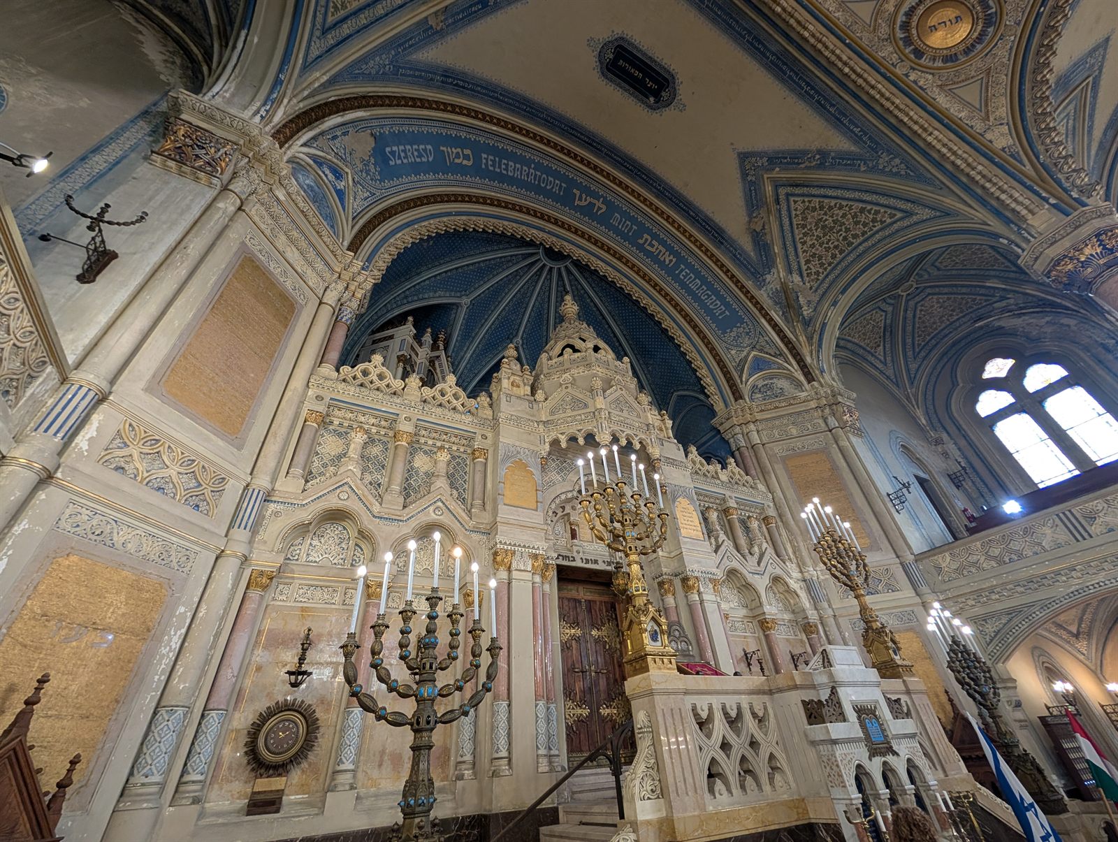 The Torah ark of the Szeged synagogue: a white marble and stone structure with a domed canopy, flanked by two tall bronze menorahs, under blue painted vaults with gilded Hebrew inscriptions